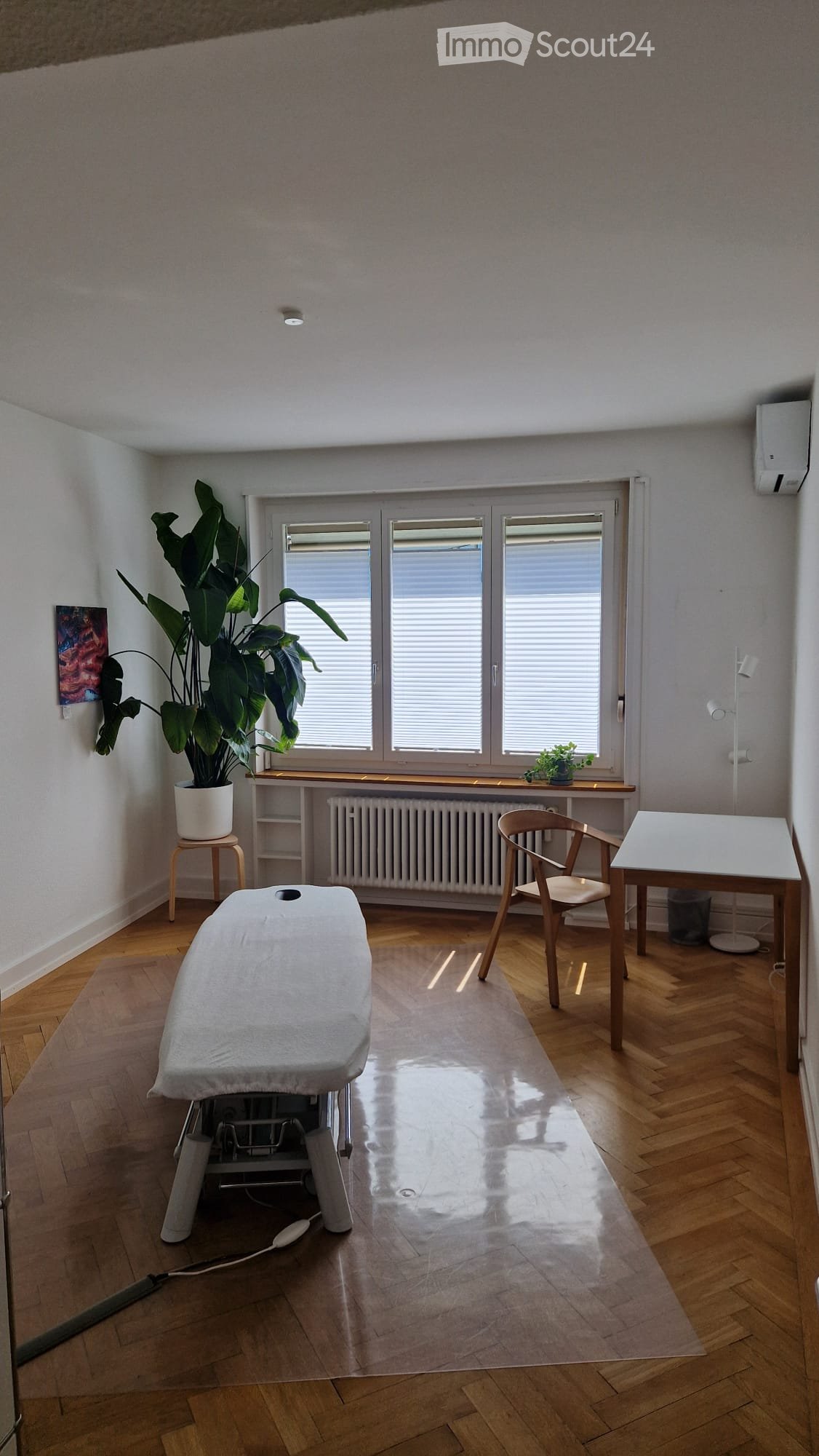 Bright room with wooden flooring, large window with blinds, a massage table, a chair, a small table, a plant on a stool, and a radiator under the window.