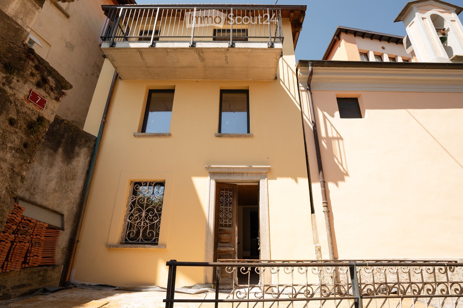 Two-story yellow building with white trim, multiple windows, and a balcony on the upper floor. The entrance features ornate metalwork and a small porch. The building is situated next to a stone wall with stacked bricks visible on the left side.