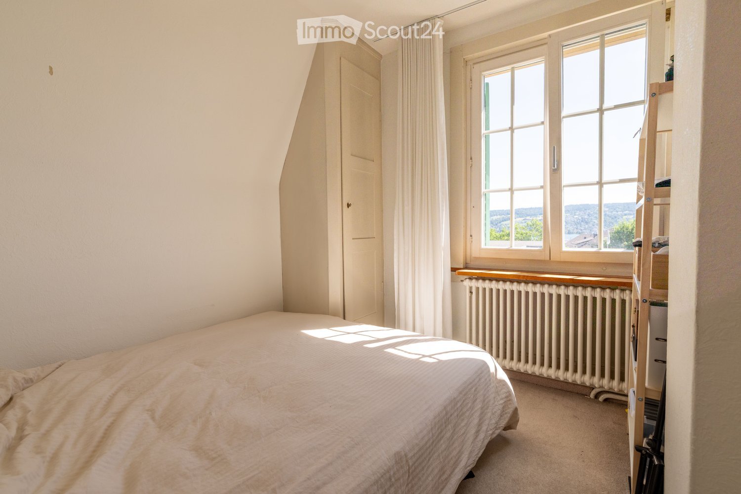 Bright bedroom with a large window offering a scenic view, featuring a neatly made bed, a radiator beneath the window, and a curtain partially drawn.