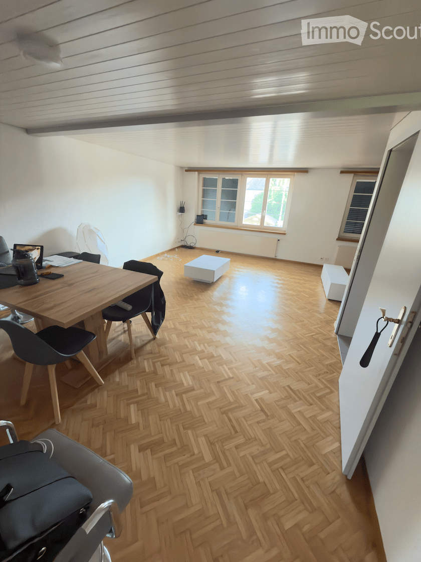 Bright room with herringbone wooden flooring, large windows, and modern furniture including a wooden table, chairs, and small white stools.