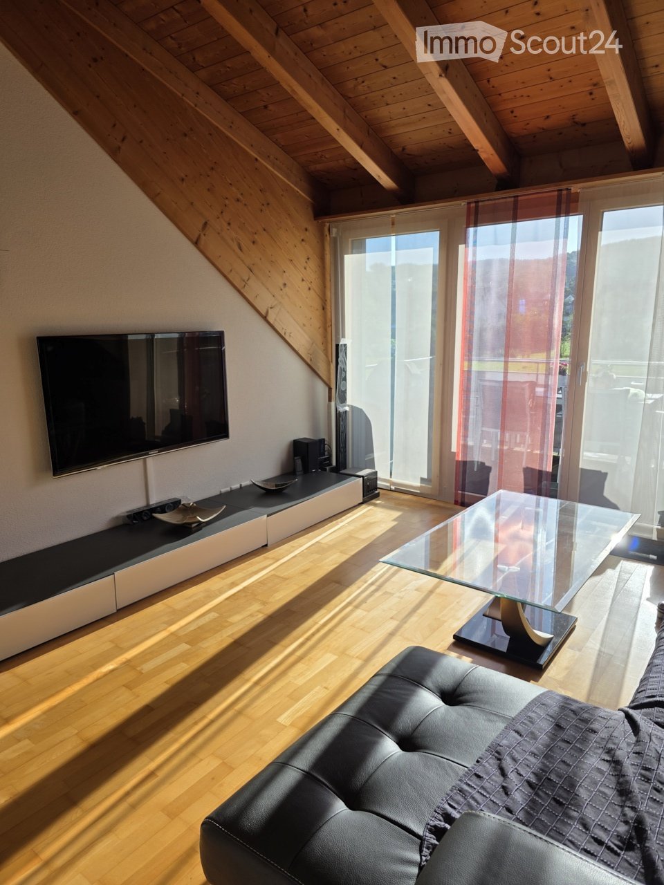 Modern living room with wooden flooring, a black leather sofa, a glass coffee table, and a flat-screen TV on a sleek entertainment unit. Large windows with sheer curtains allow natural light to fill the space.