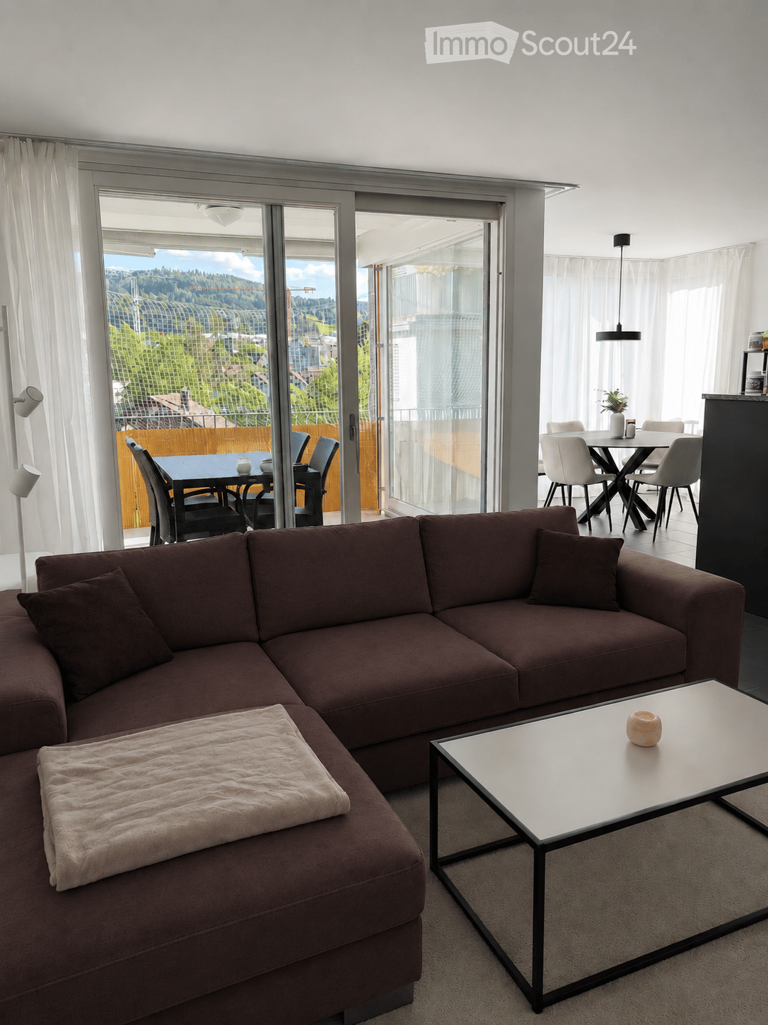 Modern living room with a brown sectional sofa, coffee table, dining area with chairs and table, large windows with a scenic view, and a hanging lamp.