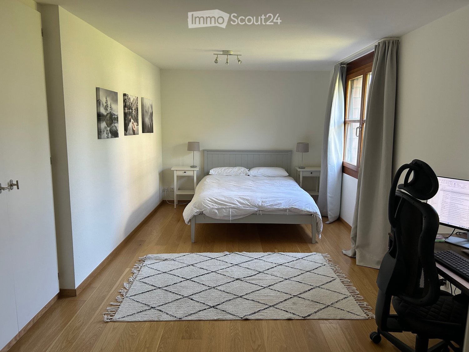 Cozy bedroom with a single bed, nightstands, lamps, a desk with a computer, and a patterned rug on the wooden floor.