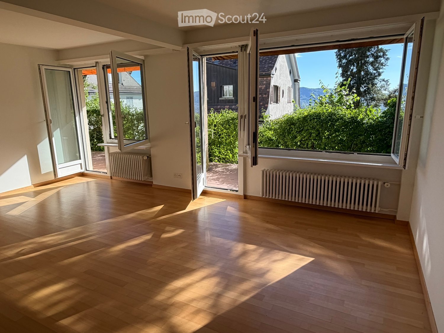 Empty room with wooden flooring, large windows, and radiators, featuring bright natural light and open views of the outdoors.