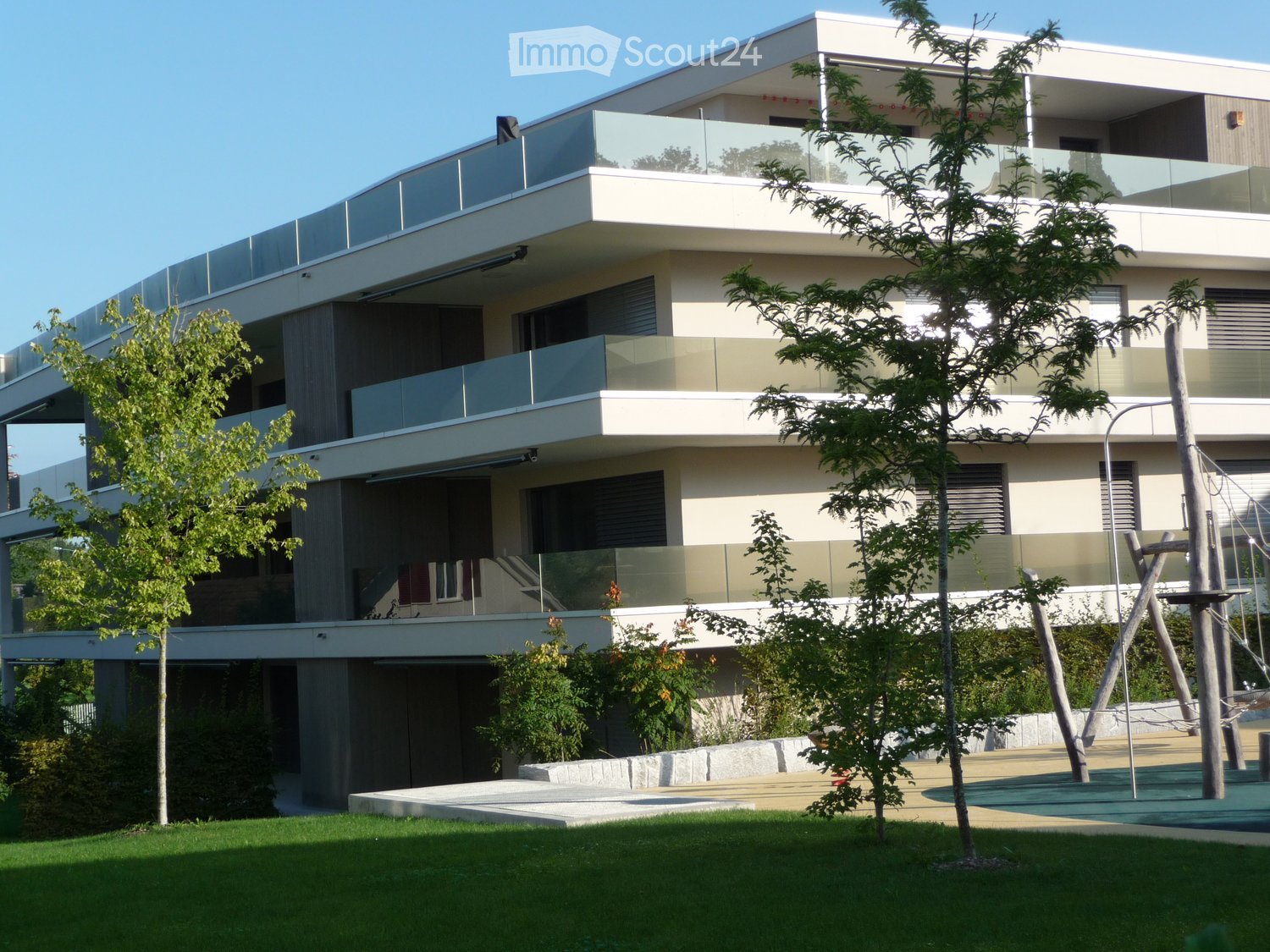 Modern two-story house with large glass balconies, clean lines, and contemporary design. The house features a raised ground floor, surrounded by a well-maintained lawn and trees.