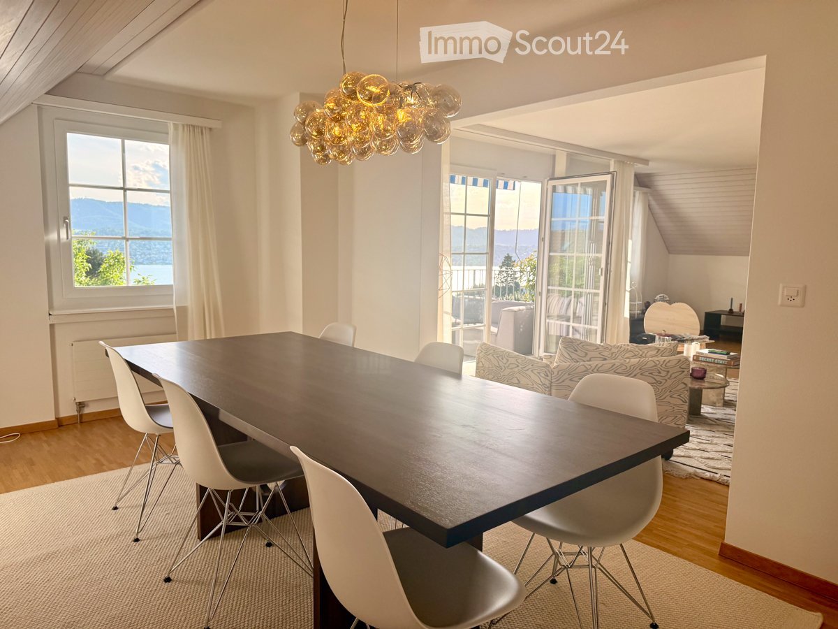 Bright dining area with a large wooden table, white chairs, and a modern chandelier. Adjacent living area features a patterned sofa, coffee table, and scenic mountain view through large windows and sliding doors.