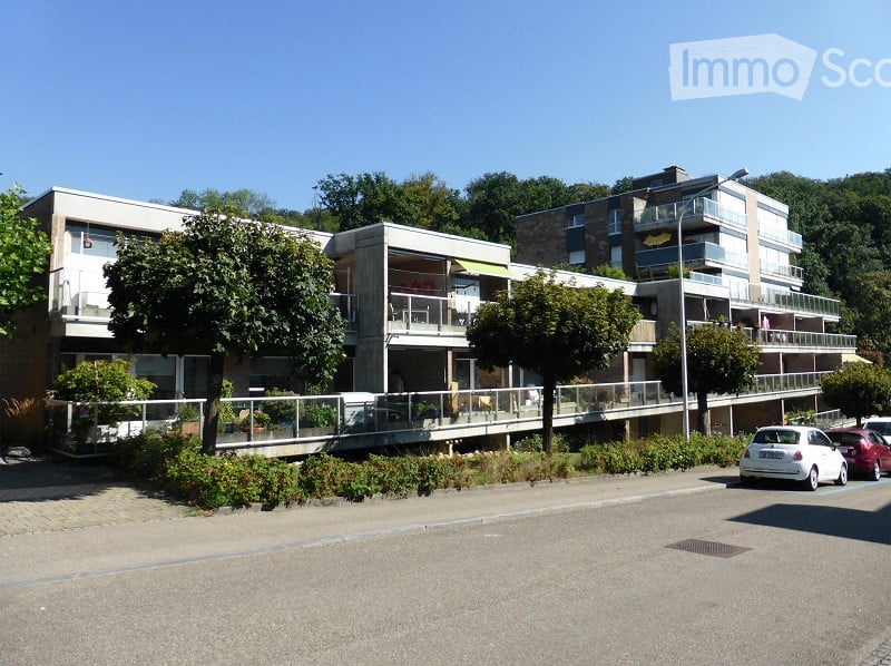 Modern multi-story residential building with balconies, surrounded by trees and parked cars.