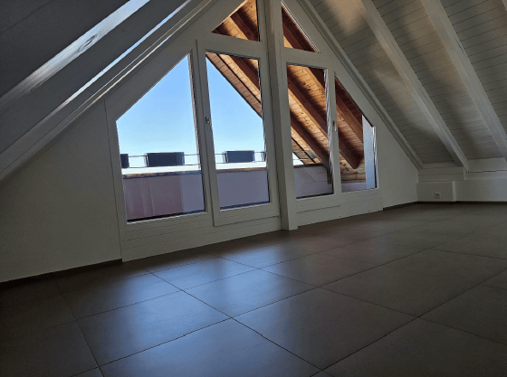 Bright, empty attic space with large skylight windows, wooden beams, and a tiled floor. Features a small balcony area with a view.