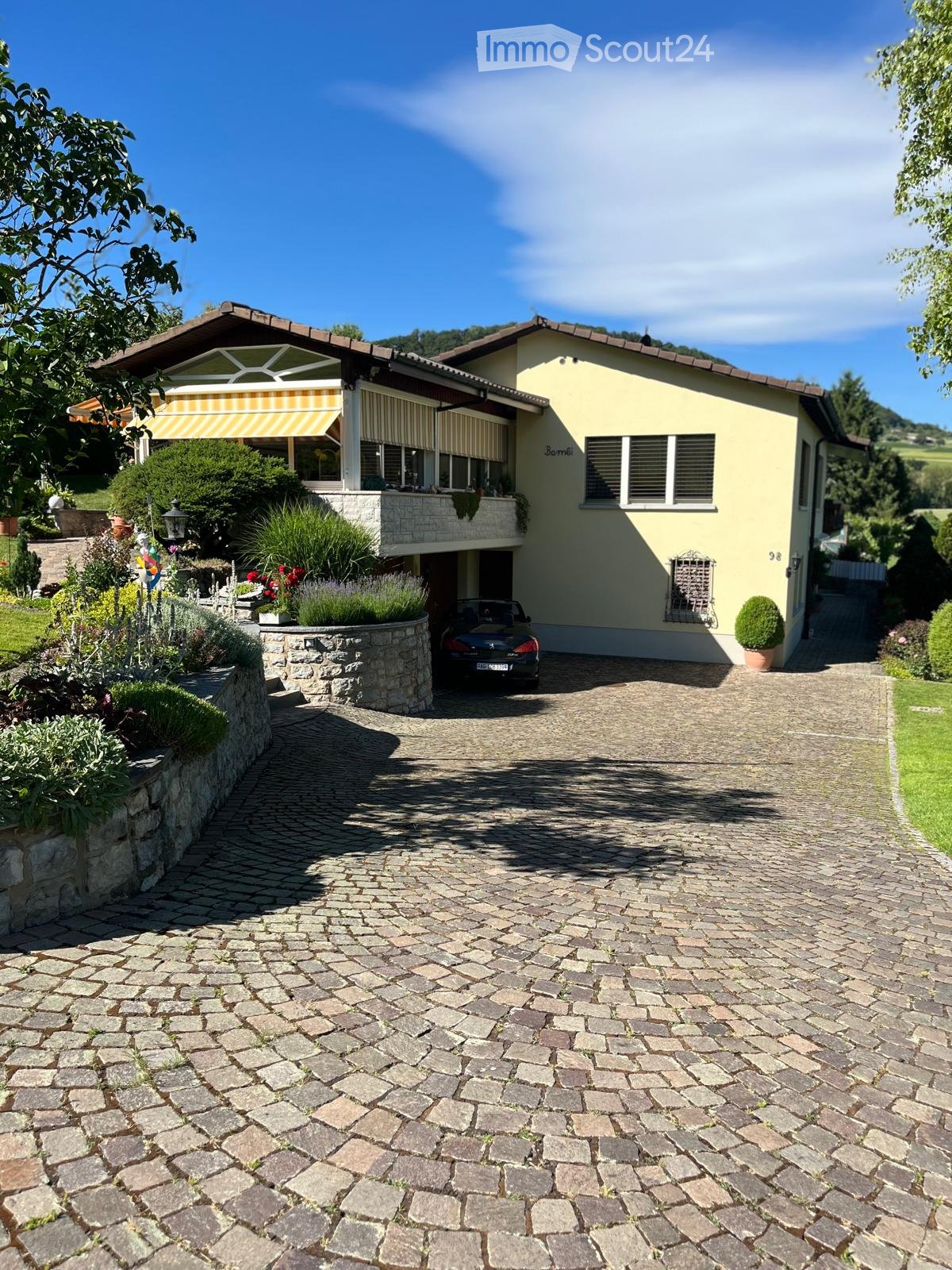 Two-story house with yellow exterior, stone driveway, garage, and garden area with plants and flowers.