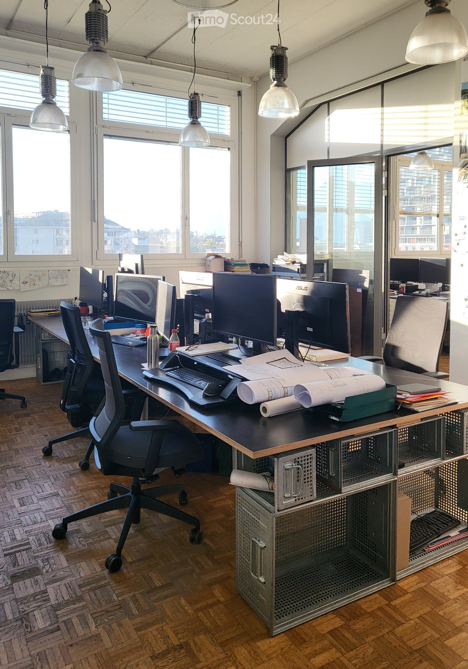 Modern office space with wooden flooring, large windows, multiple desks with computers, chairs, and office supplies.