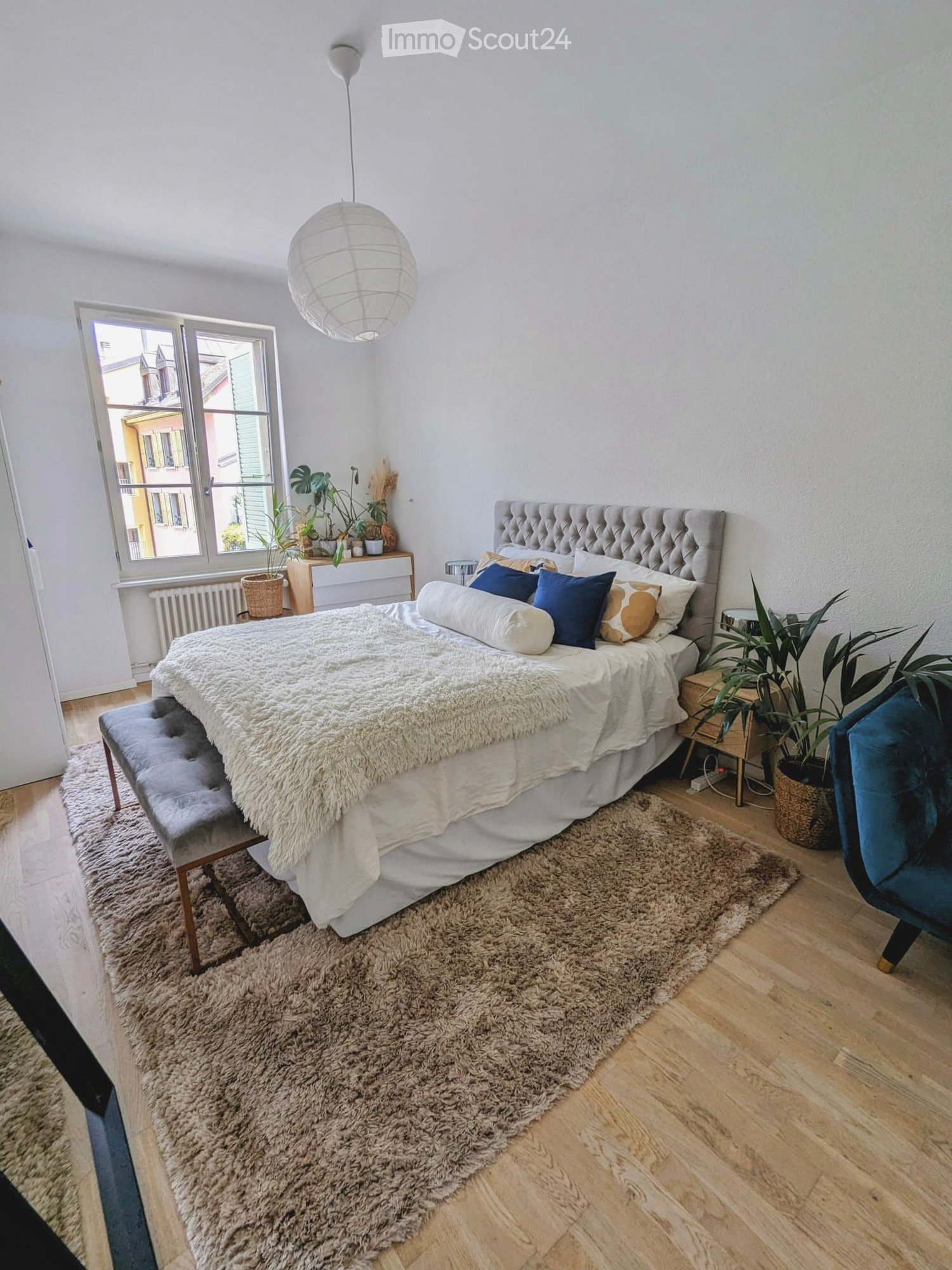 Cozy bedroom with a large bed, fluffy white bedding, blue and beige pillows, a tufted headboard, a bench at the foot of the bed, a blue armchair, a side table with potted plants, and a large shaggy rug on the wooden floor.