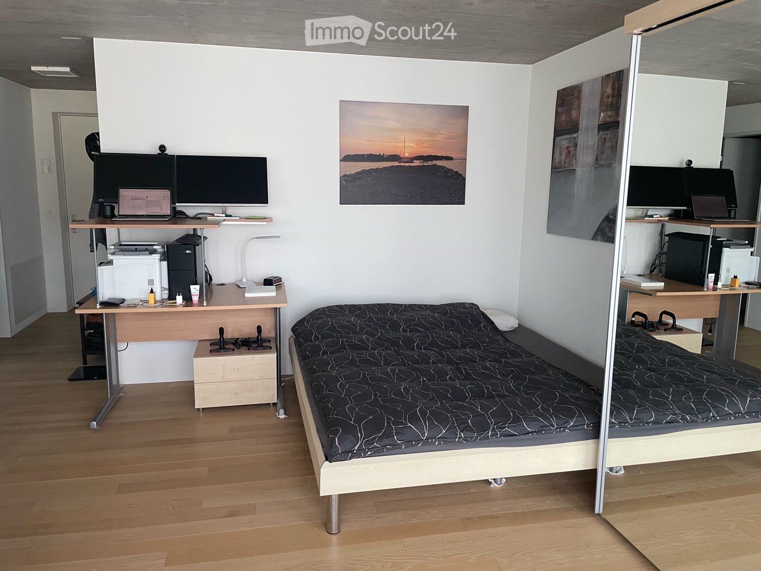 A compact, modern bedroom with a raised bed, desk, and two screens, featuring a wooden floor and white walls.