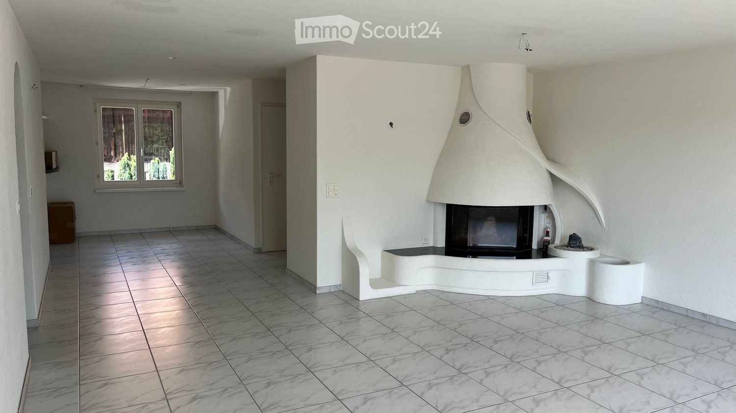 Modern living room with a white, sculptural fireplace, tiled floor, large window, and neutral-colored walls.