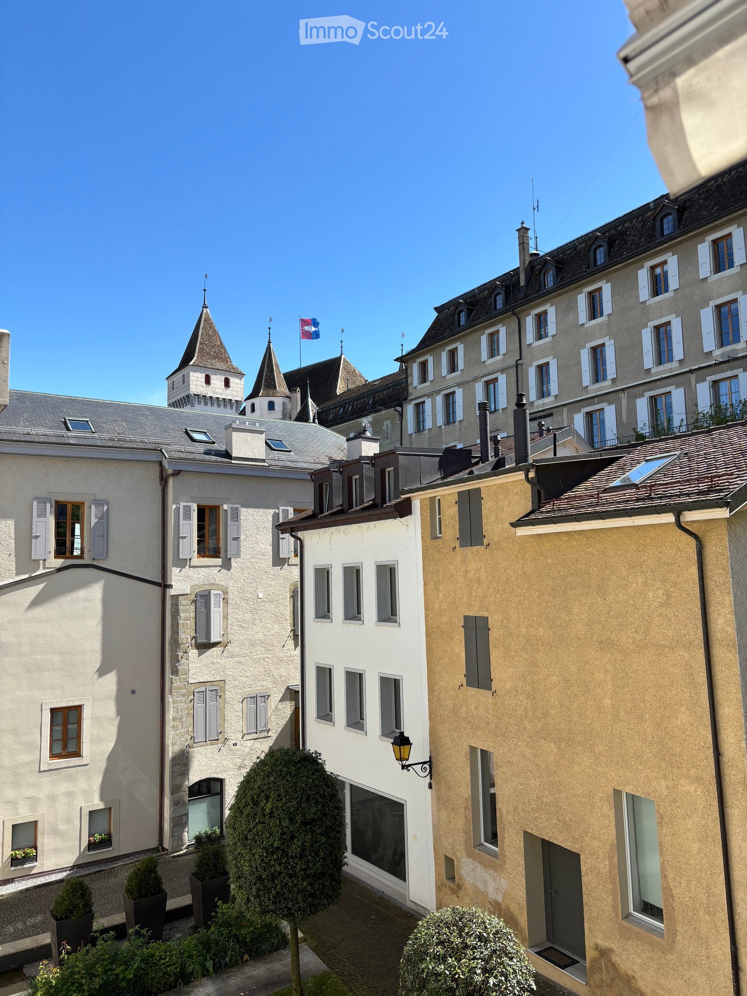 A picturesque European town scene with multi-story buildings, traditional architecture, and a clear blue sky. The buildings feature white and beige facades, shuttered windows, and ornate rooftops. A small garden area with trimmed bushes and potted plants