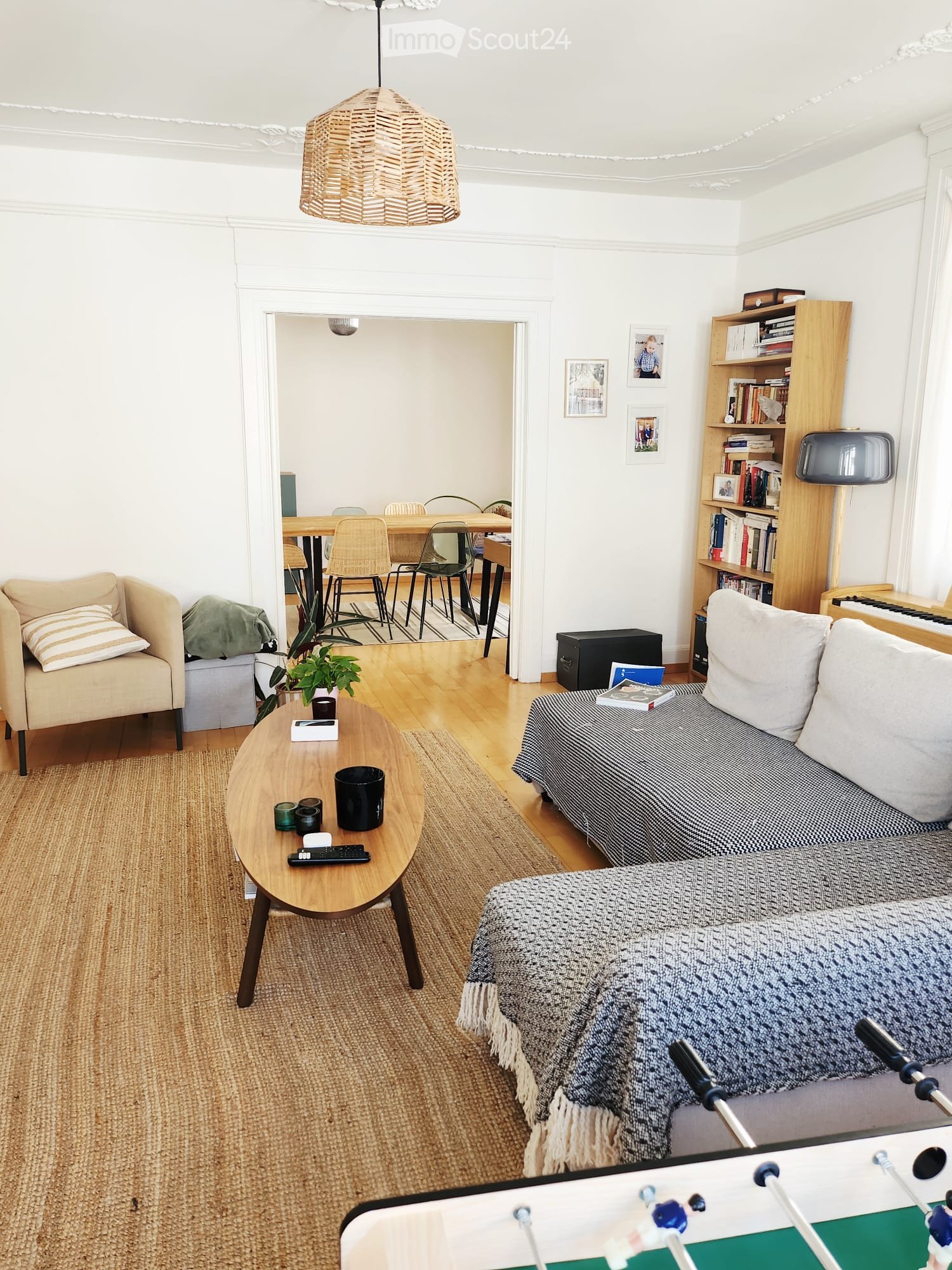 A cozy living room with a sofa, coffee table, armchair, dining area, bookshelf, and piano. The space features wooden flooring, white walls, and a hanging woven lamp.