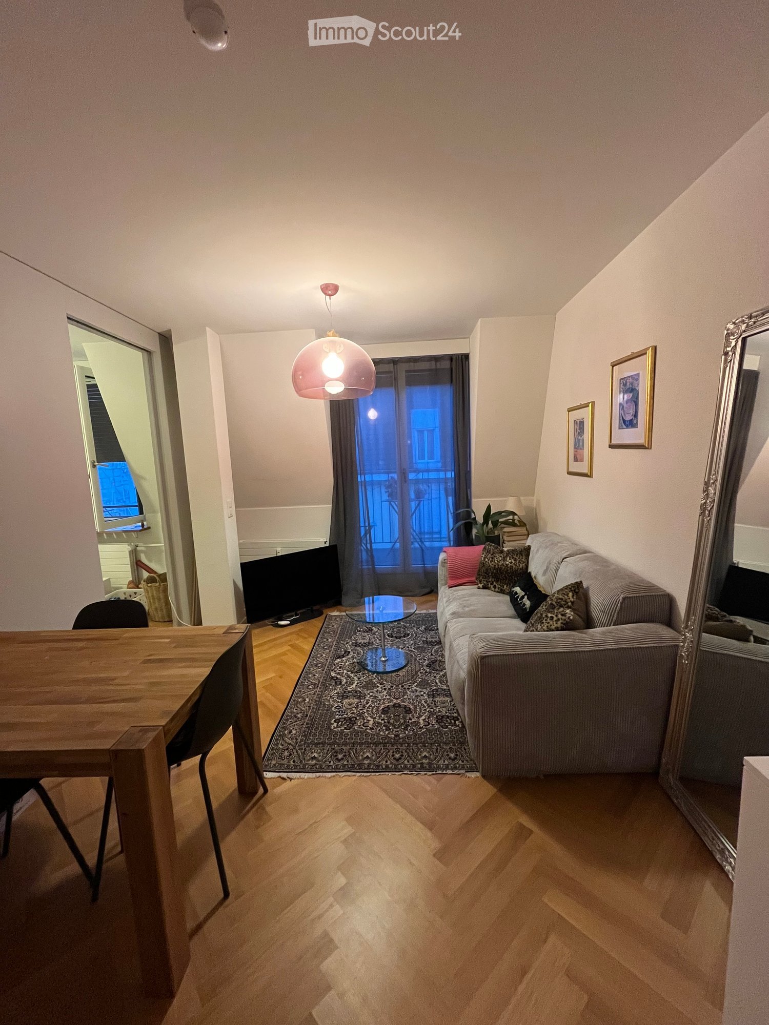 A modern living room with wooden flooring, a grey sofa with patterned cushions, a glass coffee table, a large mirror, a wooden dining table with chairs, and a television. The room features a hanging pendant light and a view of a balcony through large wind