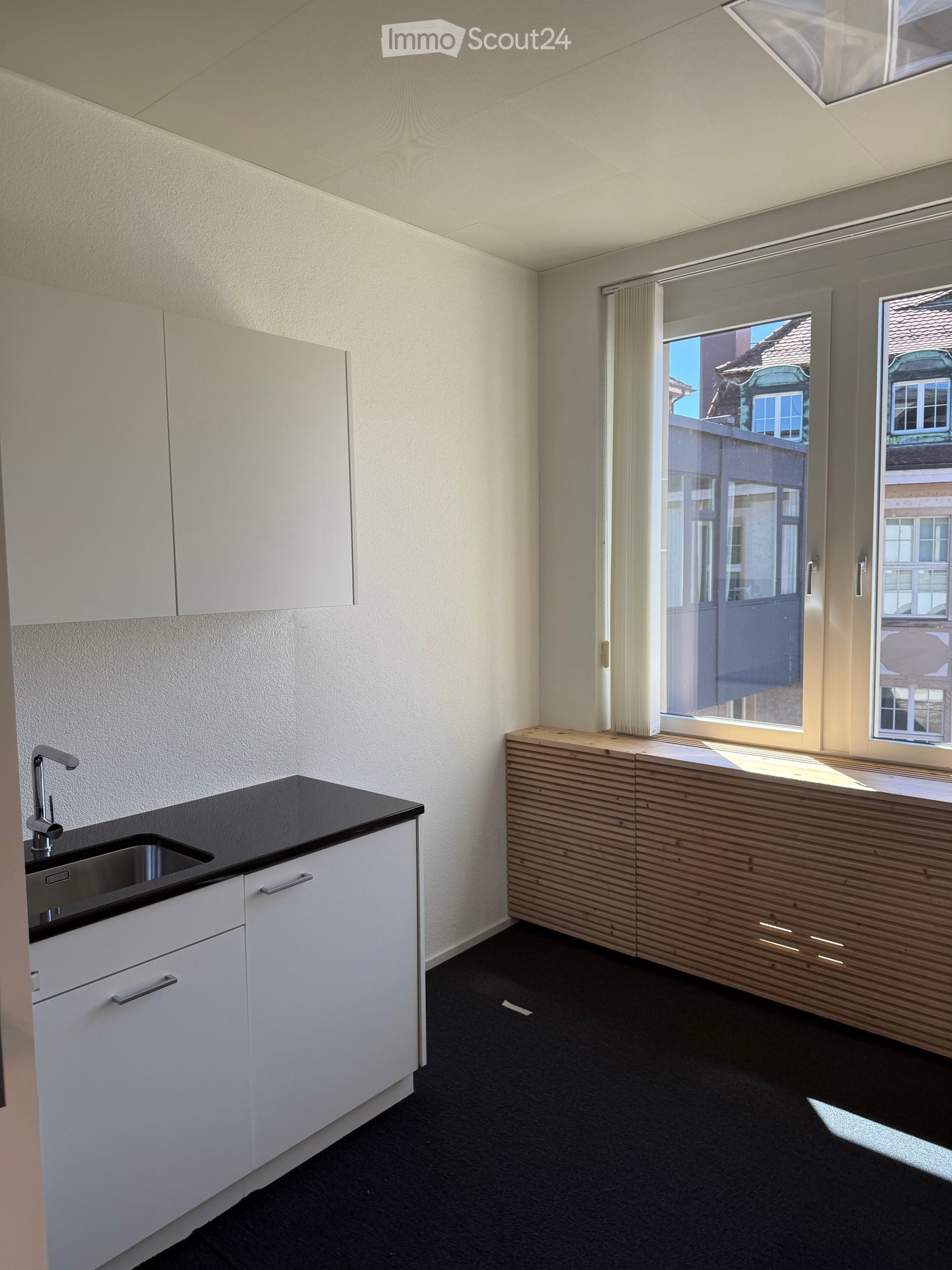 Modern kitchen with white cabinets, black countertop, and a wooden breakfast bar. Features a sink with a modern faucet and large windows offering a view of the street.