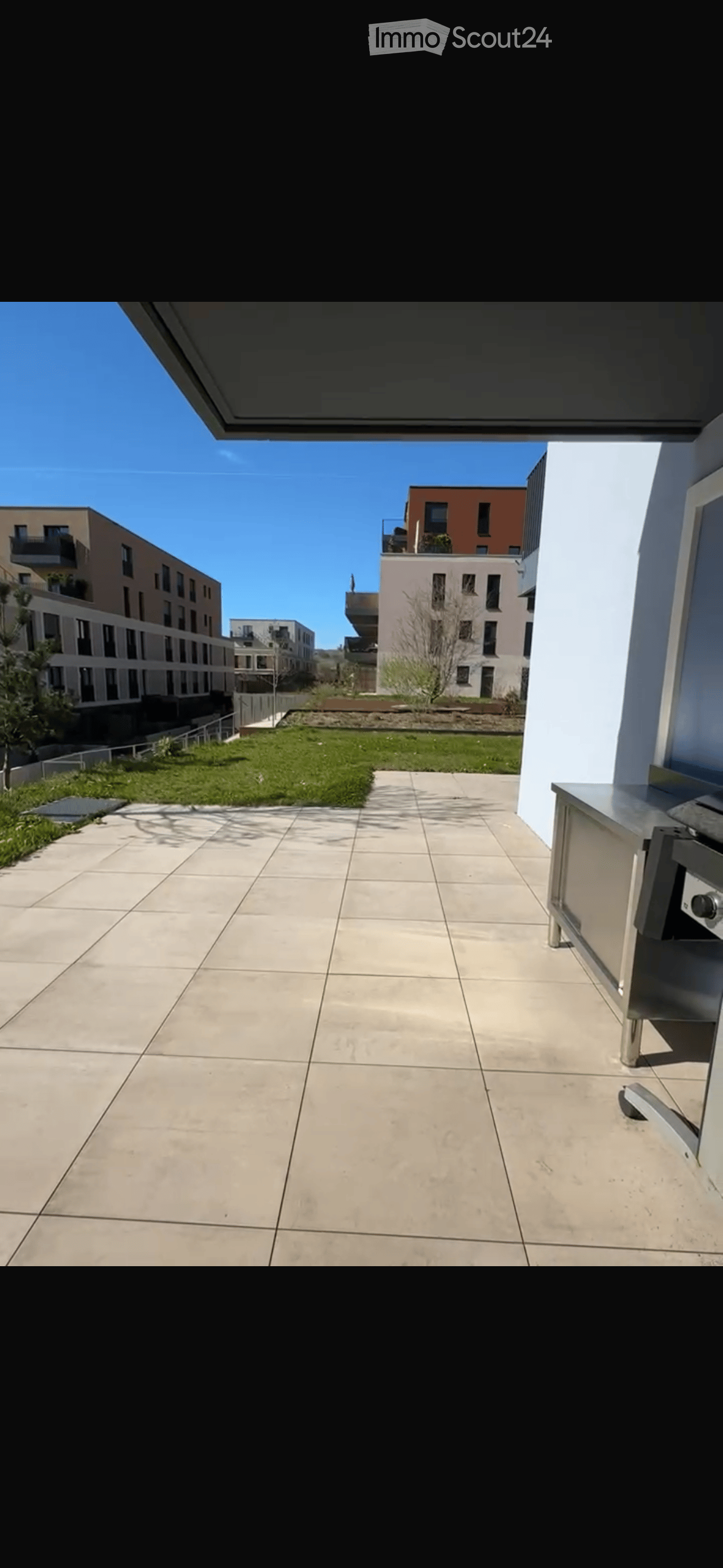 Spacious balcony with tiled floor, modern stainless steel railing, and a view of a landscaped area with grass and modern apartment buildings.