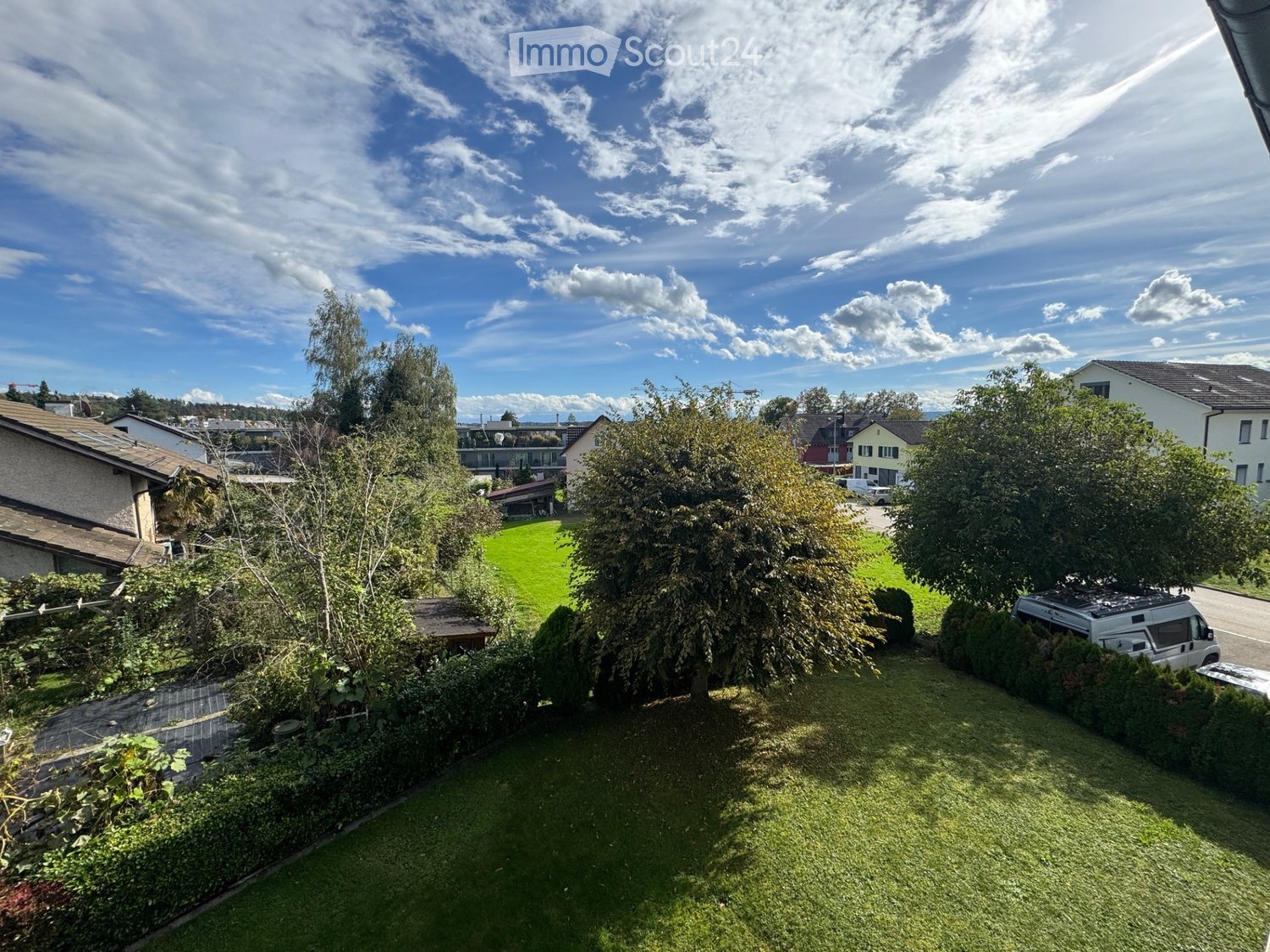 A well-maintained garden with green grass, trees, bushes, and a small wooden table. The background shows neighboring houses and a clear blue sky with scattered clouds.