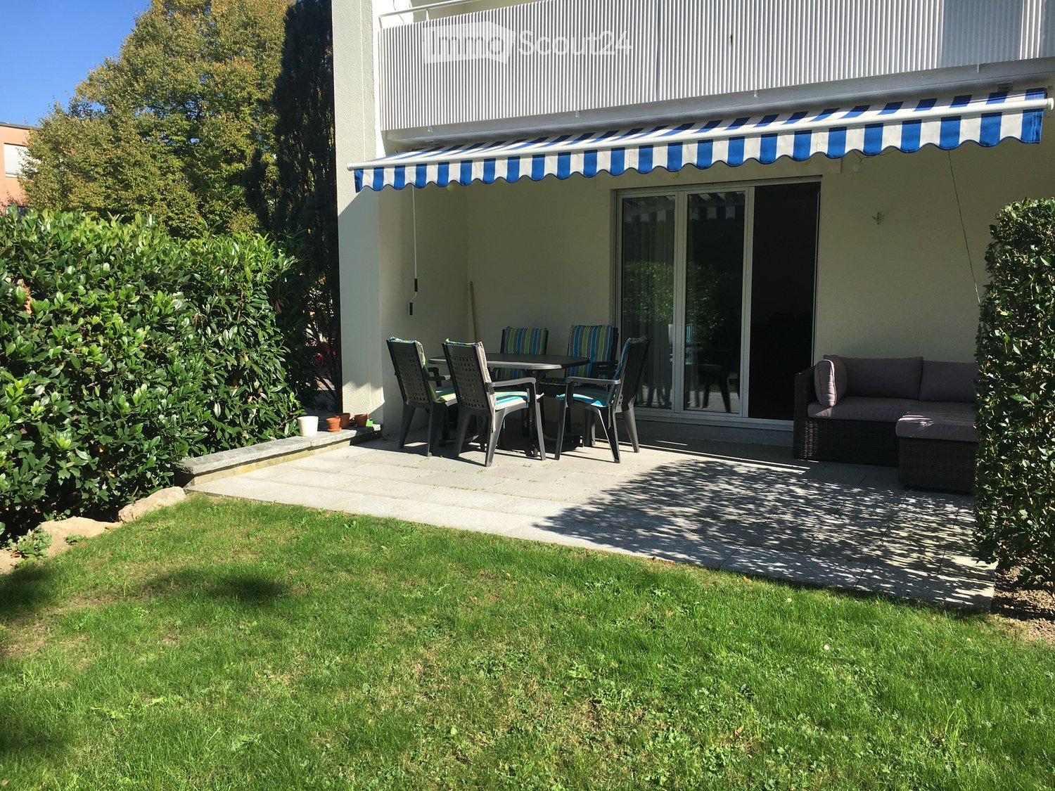 Outdoor patio area with a table, chairs, and a sofa, adjacent to a well-maintained lawn and surrounded by greenery.