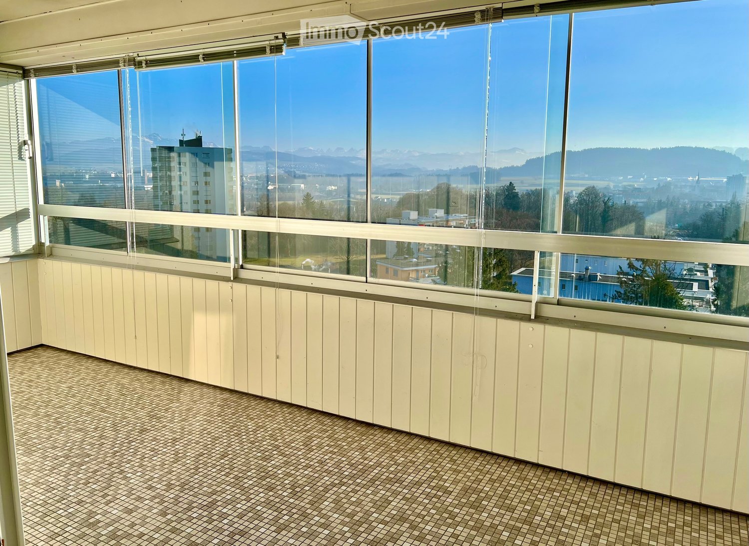 Empty room with large windows offering a scenic view of the city and mountains, featuring tiled flooring and light-colored paneling.