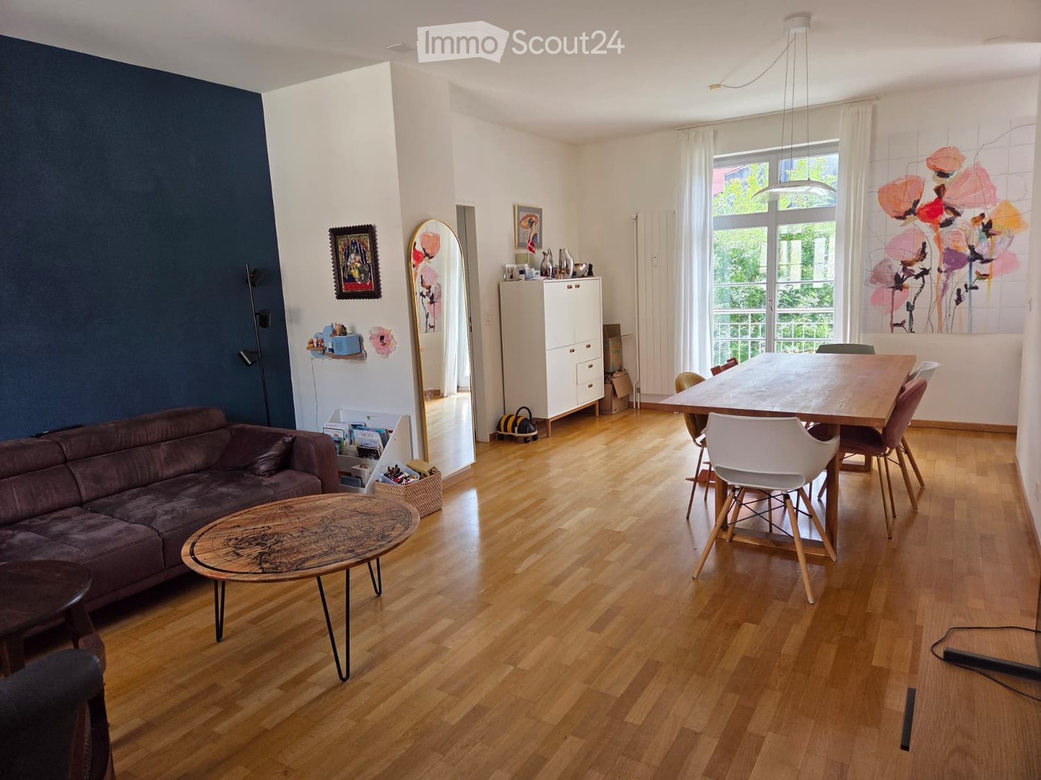 Open-plan living and dining area with wooden flooring, a dark blue accent wall, a brown leather sofa, a round wooden coffee table, a dining table with chairs, a white cabinet, and artwork on the walls.