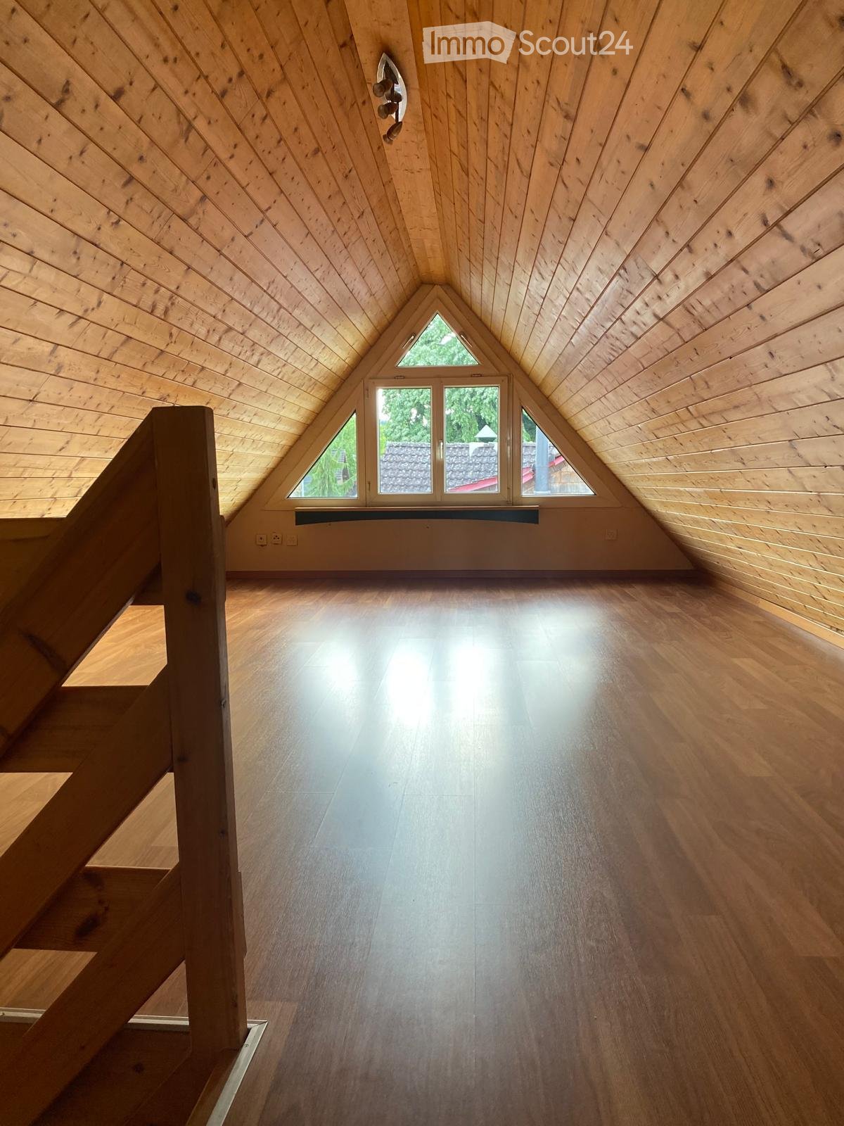 Bright, empty attic space with wooden walls, ceiling, and floor, featuring large triangular windows and a staircase leading up.