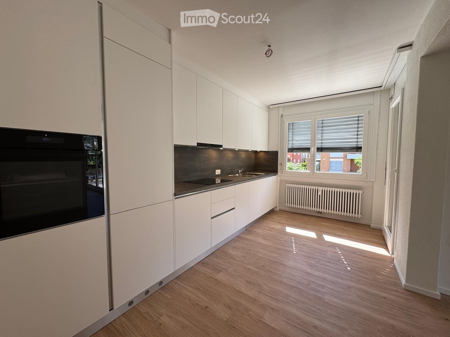 Modern kitchen with white cabinets, wooden flooring, and a black oven. Features a sink, radiator, and large windows with blinds.