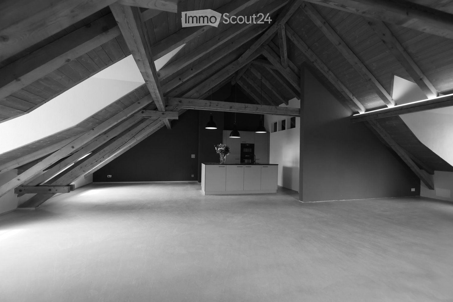 Open loft space with exposed wooden beams, concrete floor, and a small kitchen area with cabinets and a vase on the counter.