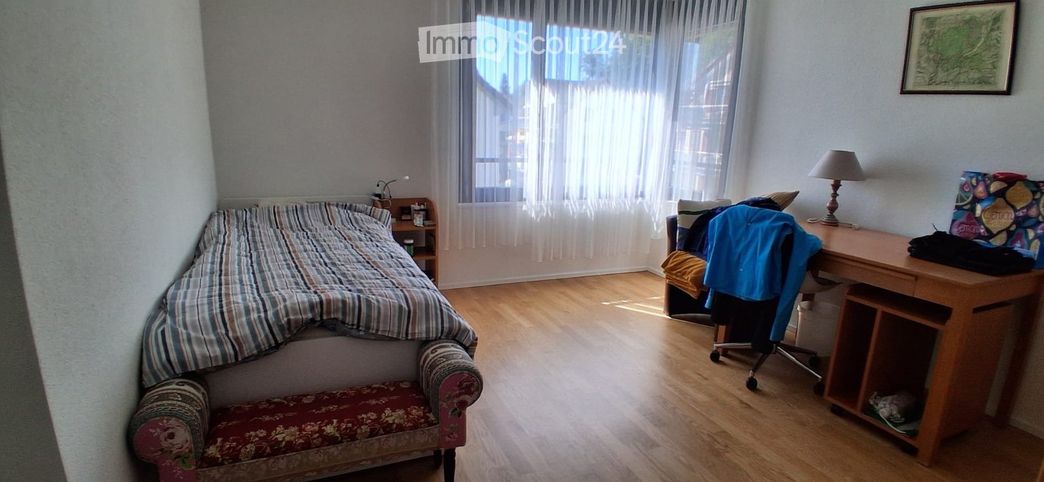 Cozy bedroom with wooden flooring, a bed with striped bedding, a small chair, a desk with a lamp, and a nightstand.