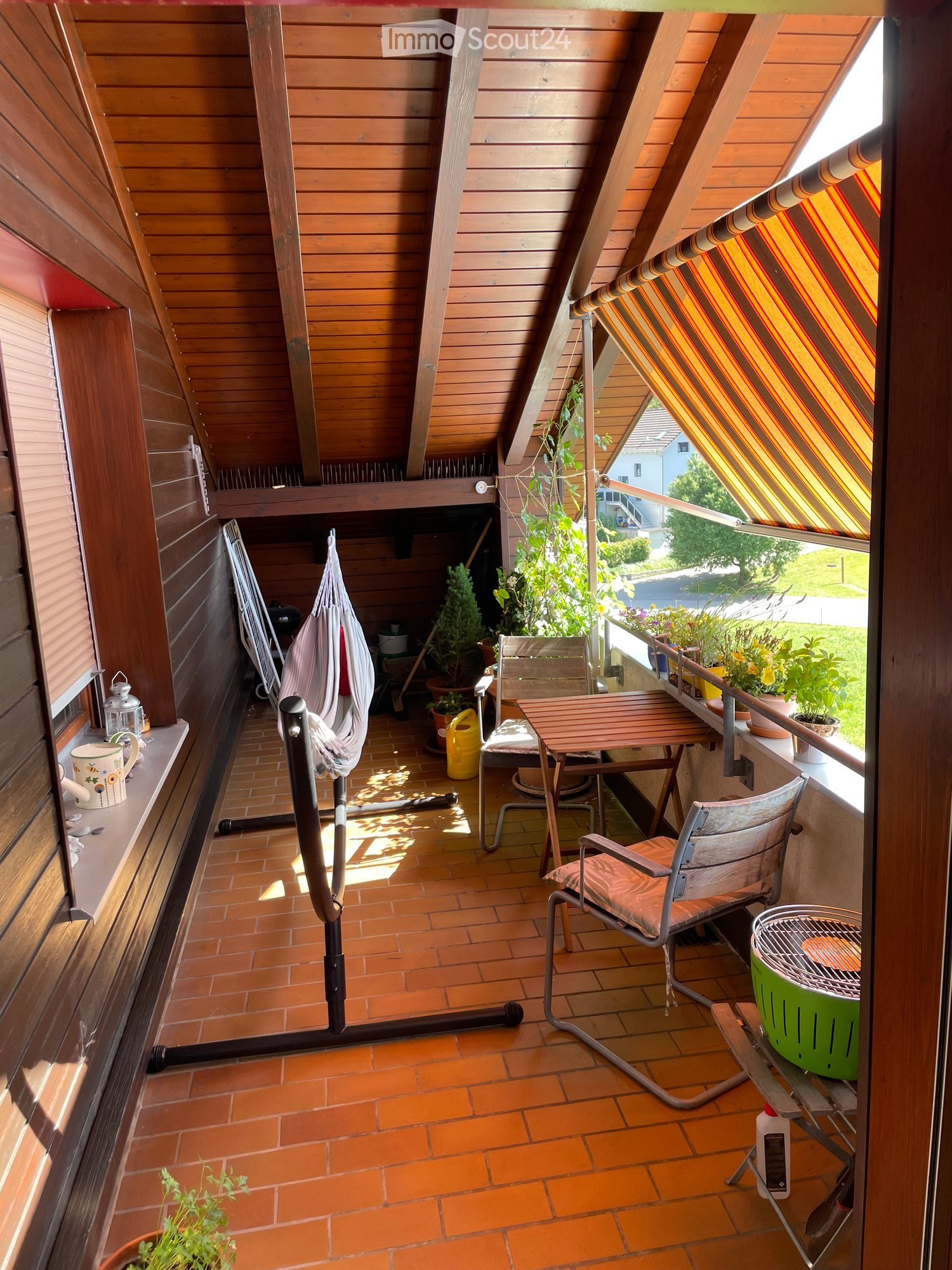 Wooden balcony with slanted roof, tiled floor, table, chairs, hammock, grill, and potted plants.