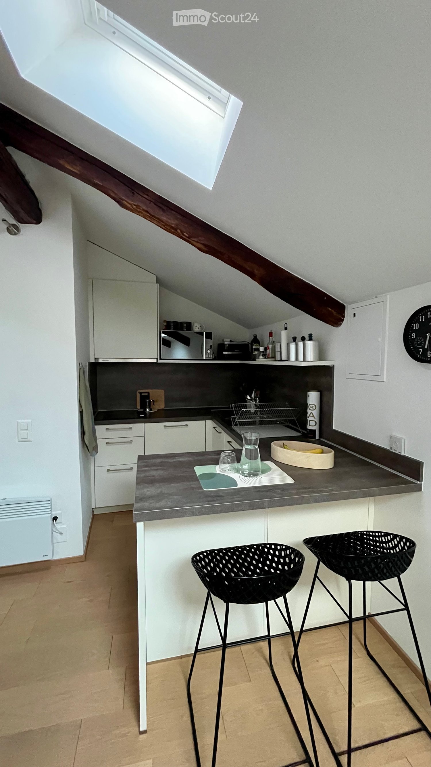 Modern kitchen with white cabinets, dark countertops, and two black stools at the island. Features a sink, microwave, and various small appliances on the counter.