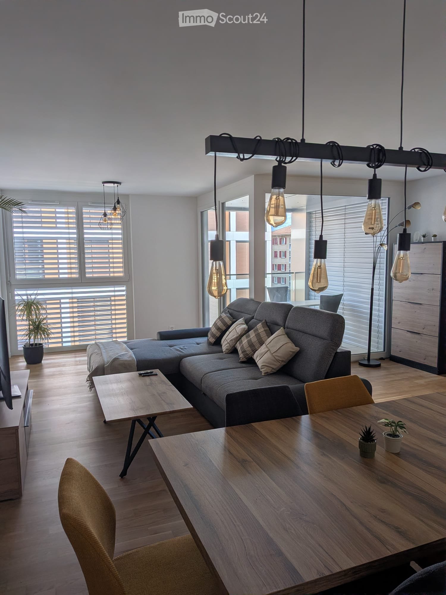 Modern living room with grey sectional sofa, wooden coffee table, dining table, chairs, and pendant lights.