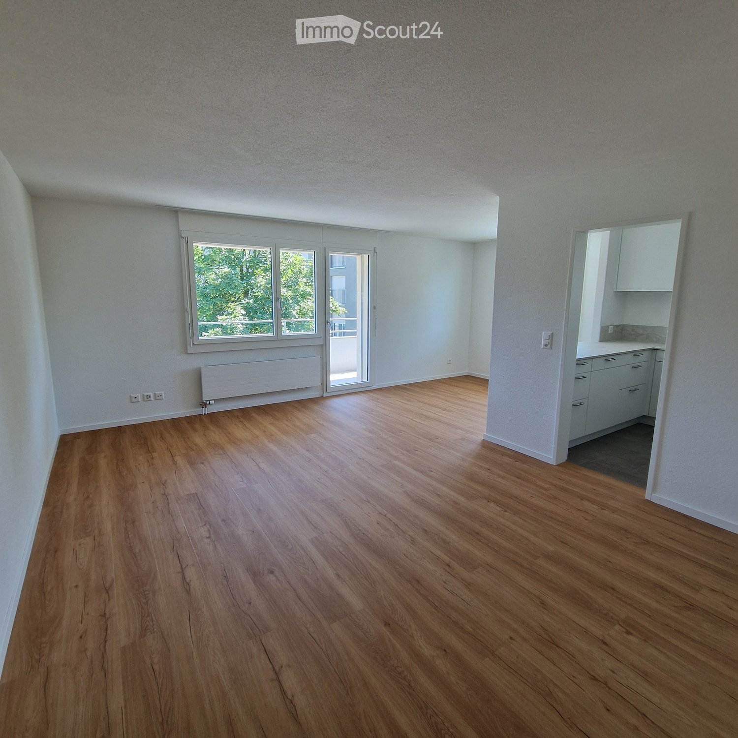 Empty room with wooden flooring, white walls, large windows, and a radiator. The room is unfurnished and has a door leading to another area.
