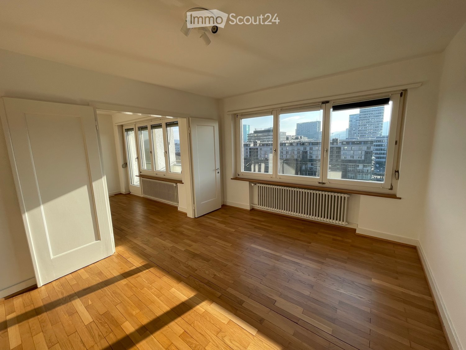 Empty room with wooden flooring, white walls, large windows with city view, radiators, and a ceiling light fixture.