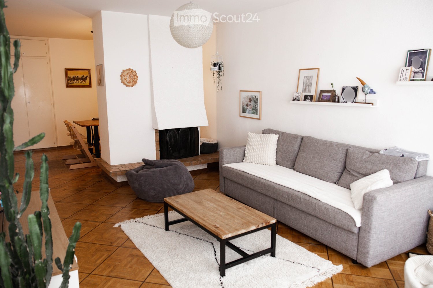 Living room with grey sectional sofa, wooden coffee table, beige rug, fireplace, wall-mounted shelf, picture frames, and a small table with chairs in the background.