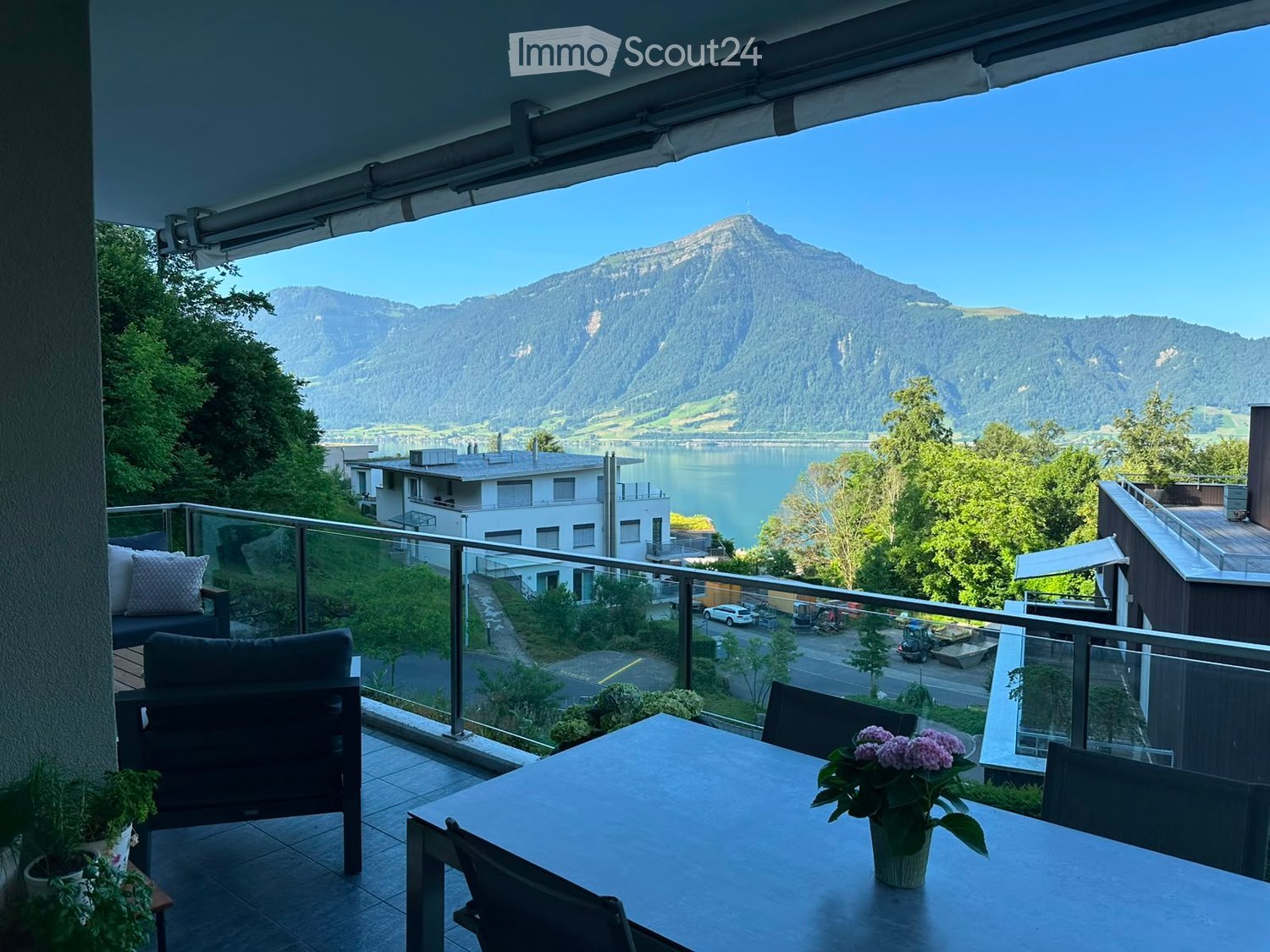 Balcony with a scenic mountain and lake view, furnished with a table, chairs, sofa, and flower pot.