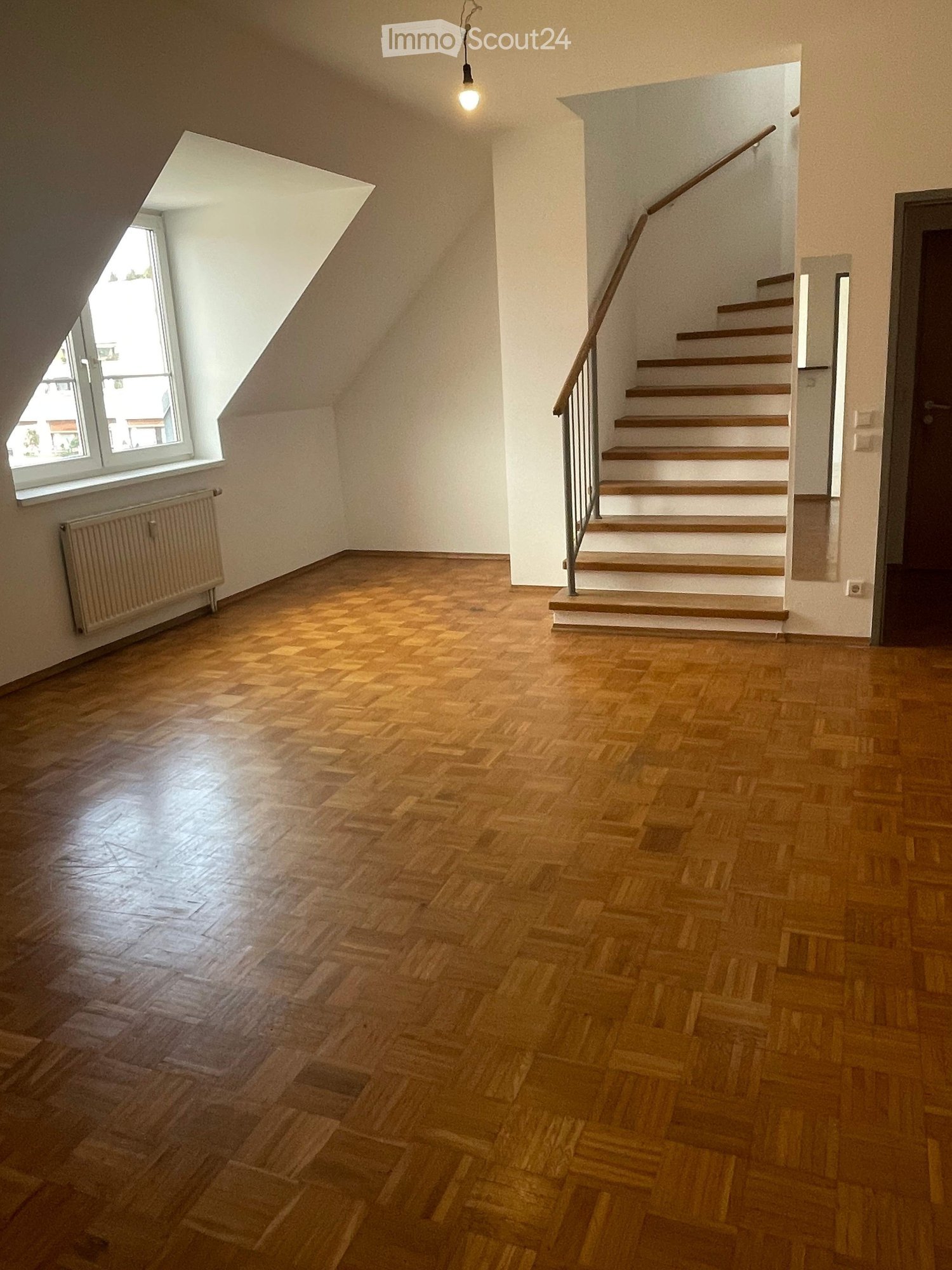 Empty room with wooden flooring, a staircase leading upwards, a window with a radiator below, and a hanging light fixture.