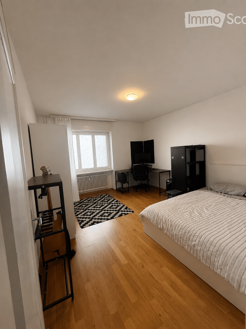 Bright bedroom with wooden flooring, large window with curtains, bed, desk with chair and screen, cabinet, mirror, and small table with items.