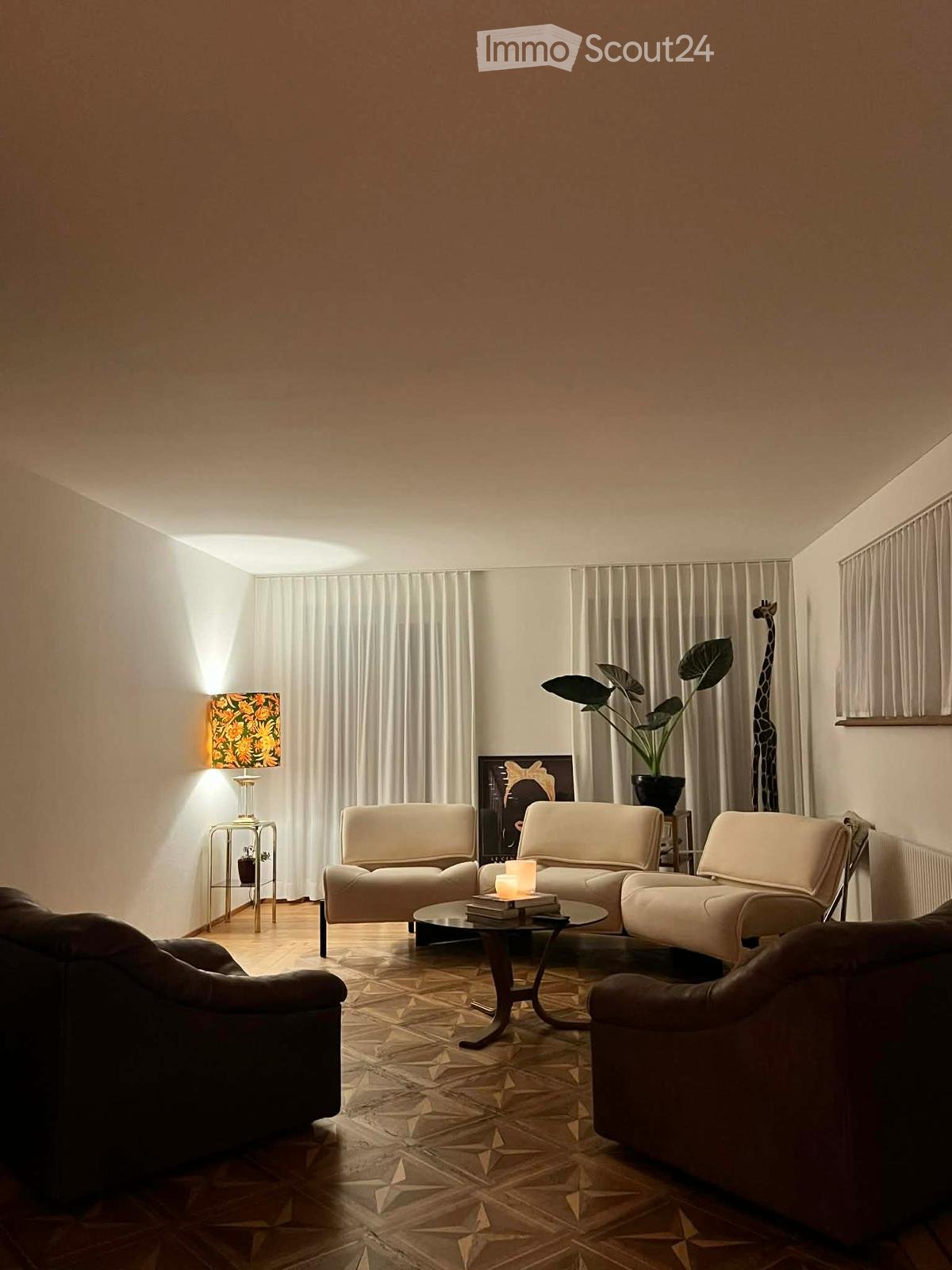 Modern living room with a cream-colored sectional sofa, a dark brown armchair, a glass coffee table, a decorative lamp, and a plant in a pot. The room features patterned tile flooring and large windows with white curtains.