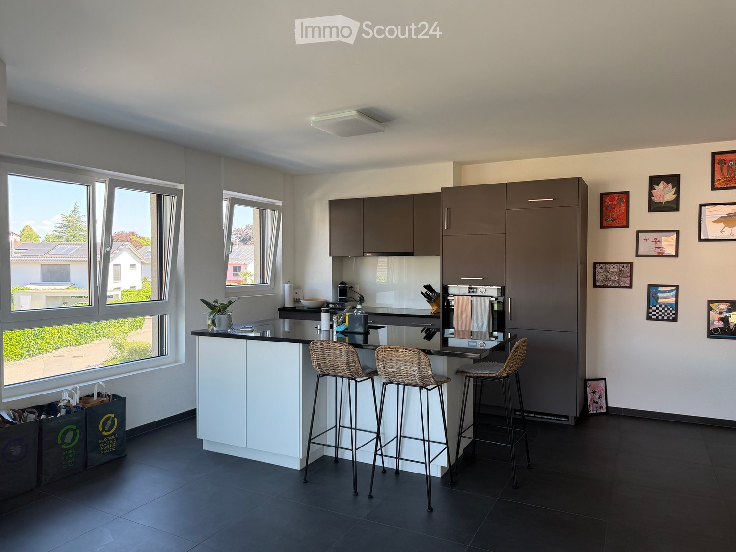 Modern kitchen with grey cabinets, white island, black countertop, three bar stools, and various appliances. Large windows provide natural light, and framed pictures decorate the wall.