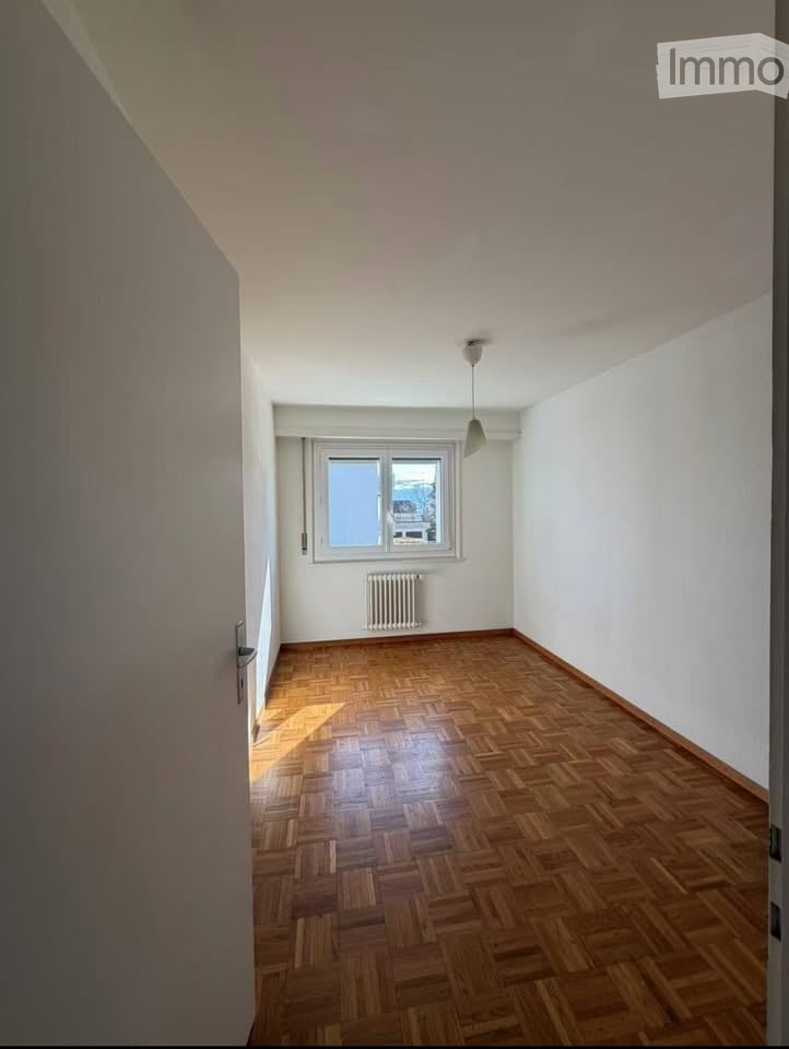 Empty room with wooden parquet flooring, white walls, a single window with a radiator below, and a ceiling light fixture.