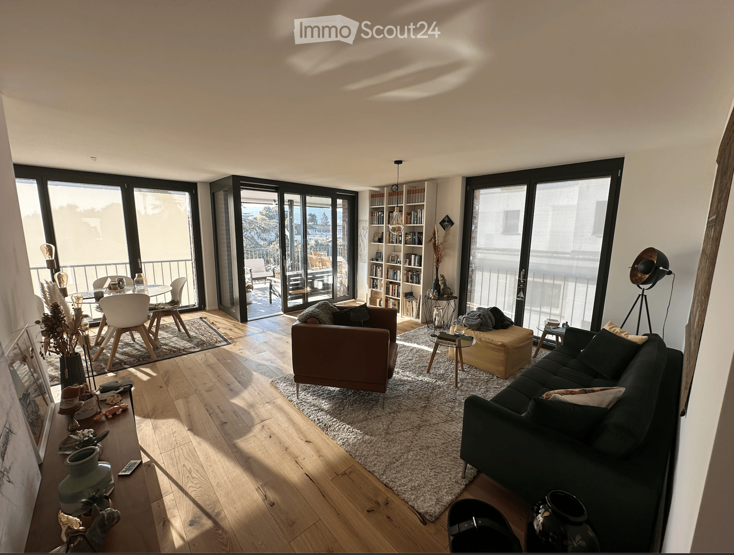 Modern living room with large windows and balcony access, featuring a dark grey sofa, brown armchair, coffee table, dining area with chairs, bookshelf, and decorative items.