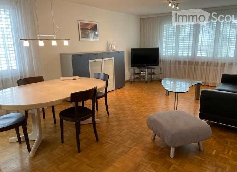 Modern furnished room with hardwood flooring, a round dining table with chairs, a sofa with an ottoman, a television on a stand, and a cabinet with a picture frame on top.
