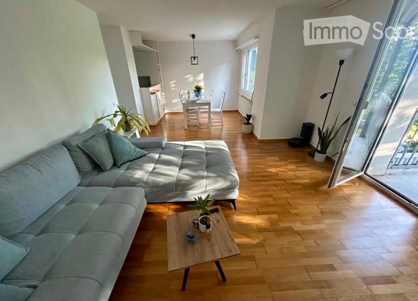 Bright living room with light grey sectional sofa, wooden coffee table, dining area with white table and chairs, and several potted plants. Natural light streams in from large windows and a glass door leading to a balcony.