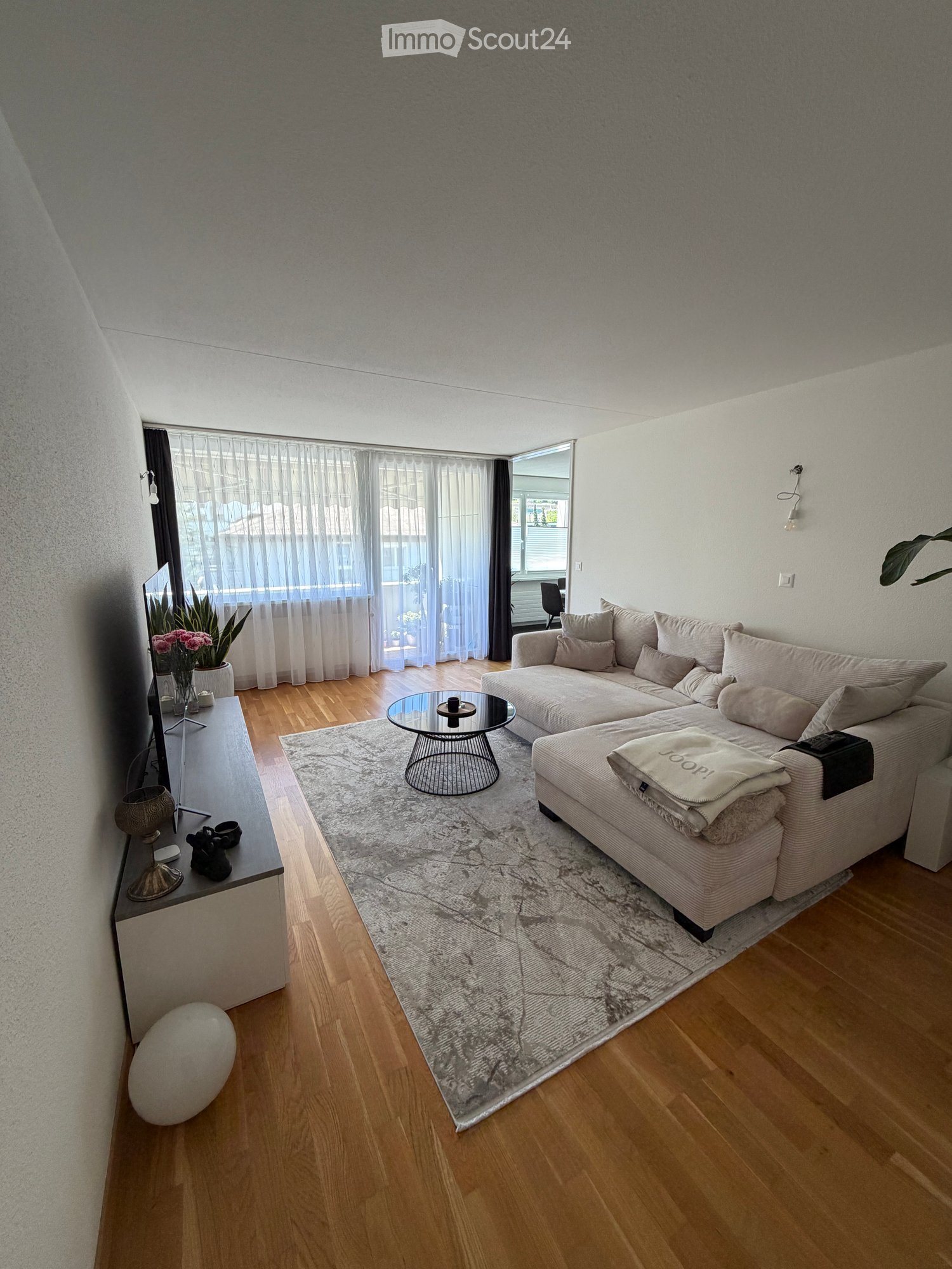 Modern living room with a sectional sofa, coffee table, area rug, and decorative items including a large white ball and a vase with flowers. Large windows with sheer curtains let in natural light.