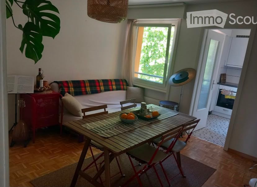 A cozy living and dining area with a wooden dining table, chairs, a sofa with a plaid throw, a bowl of fruit, and a hanging lamp. The room features wooden flooring, large windows with curtains, and a view of greenery outside.