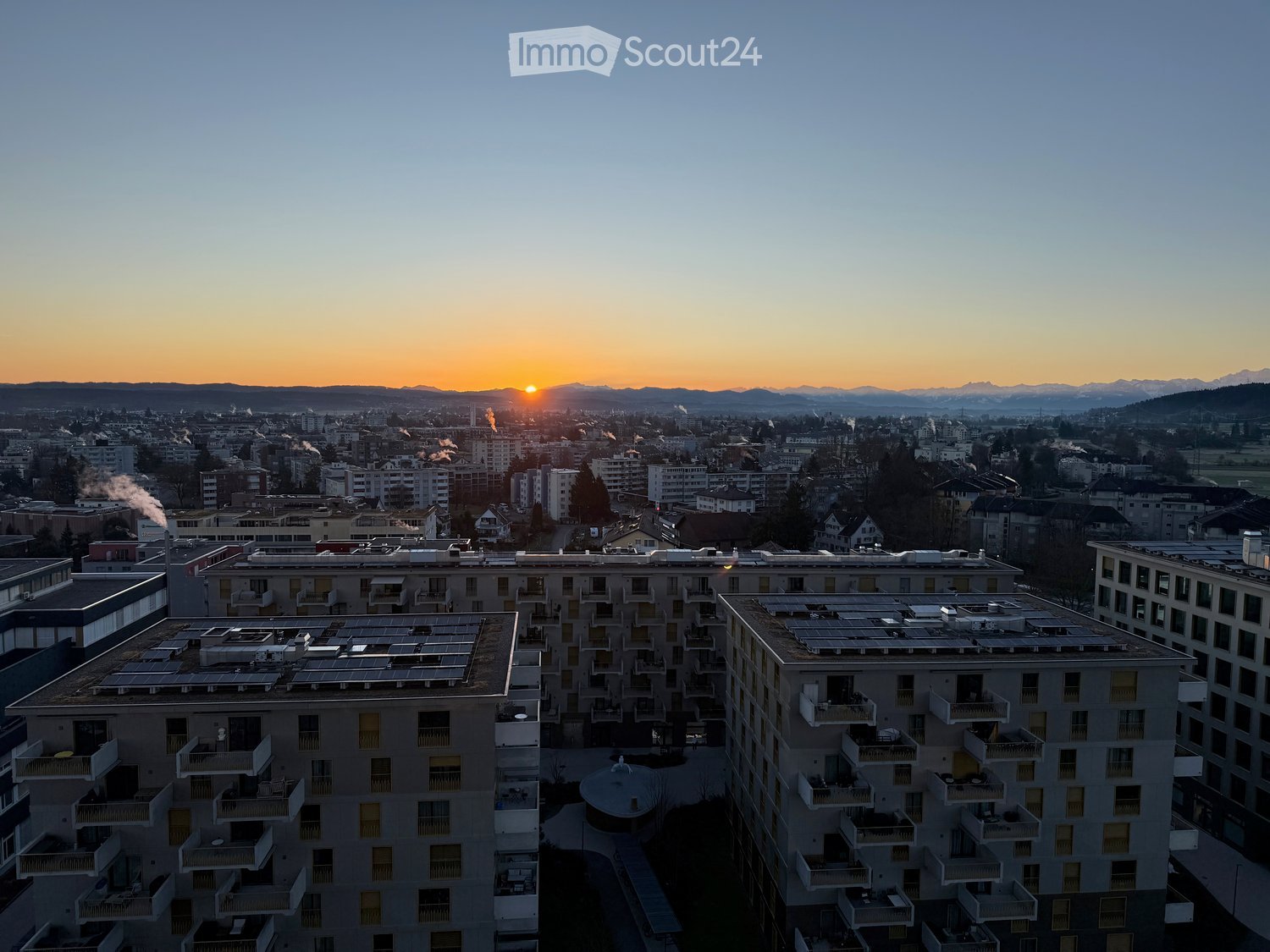 A modern apartment complex with multiple buildings, balconies, and solar panels on the rooftops, set against a cityscape with a sunset view of mountains in the background.