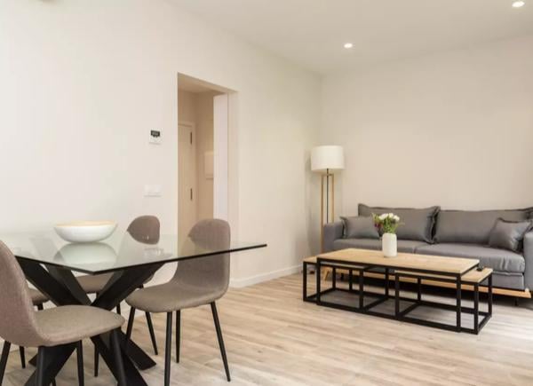 Modern, minimalist living and dining area with light grey sofa, coffee table, glass dining table, and four chairs. The space features light wooden flooring and white walls.