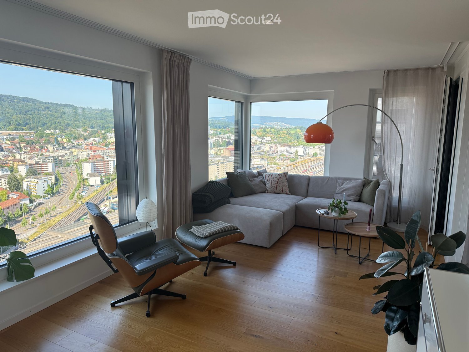 Modern living room with large windows offering a scenic city view, featuring a grey sectional sofa, a leather armchair, a small round table, a floor lamp, and a potted plant.
